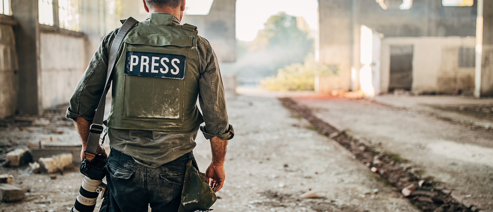 Man holding camera with a vest marked "press" walks in a damaged road full of rubble.