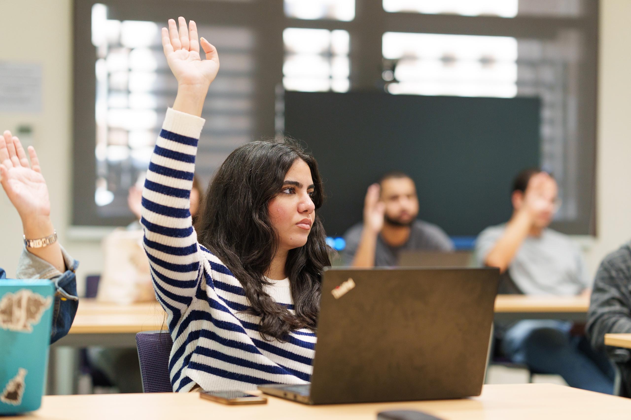 a girl with long black hair wearing a white and black shirt and raising her hand in a classroom