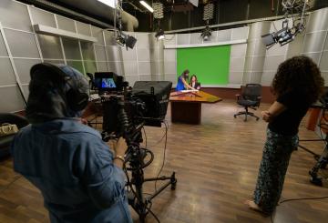 students working in a studio behind the camera filming a news anchor