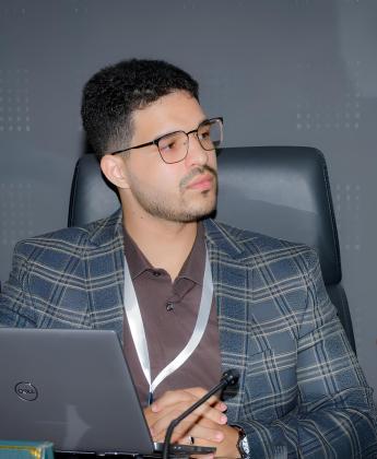 A man wearing glasses and a plaid blazer sits at a desk with a laptop and microphone, looking to the side as if listening attentively during a meeting or discussion.