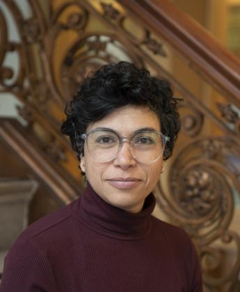 a person with short curly dark hair and glasses, wearing a maroon turtleneck, standing in front of an ornate wooden staircase with carved details, looking directly at the camera.