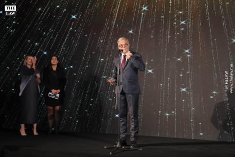 A man with a navy suit and a red tie stands in front of a conference background at a microphone, holding a glass award in his hand