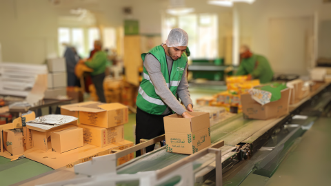 Man wearing green vest and hairnet packs a box with food supplies in a warehoues