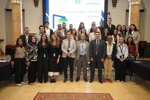 roundtable event attendees standing in rows inside a conference hall, dressed in business attire, with a presentation screen displaying Arabic text in the background.