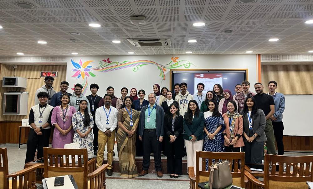 A large group of around 30&ndash;35 people stand together in a lecture hall, posing for a group photo after an academic event. The participants, of diverse genders and backgrounds, are arranged in rows facing the camera, many wearing name badges. Behind them is a presentation screen and a colorful mural on the wall, with rows of wooden chairs visible in the foreground.