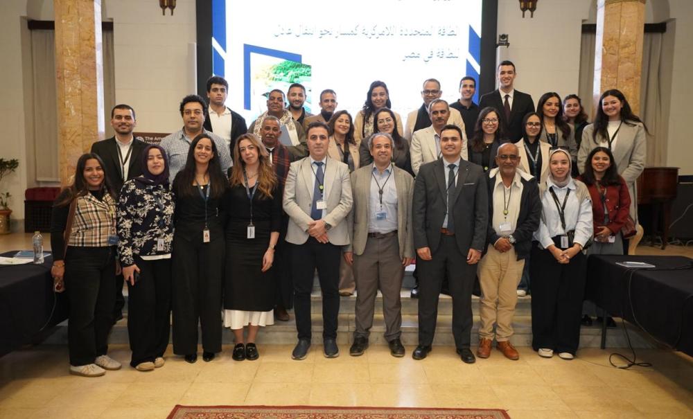 roundtable event attendees standing in rows inside a conference hall, dressed in business attire, with a presentation screen displaying Arabic text in the background.