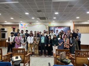 A large group of around 30&ndash;35 people stand together in a lecture hall, posing for a group photo after an academic event. The participants, of diverse genders and backgrounds, are arranged in rows facing the camera, many wearing name badges. Behind them is a presentation screen and a colorful mural on the wall, with rows of wooden chairs visible in the foreground.