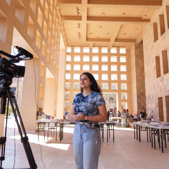 A student stands on campus speaking to a camera set on a tripod, with tables and palm trees in the background.